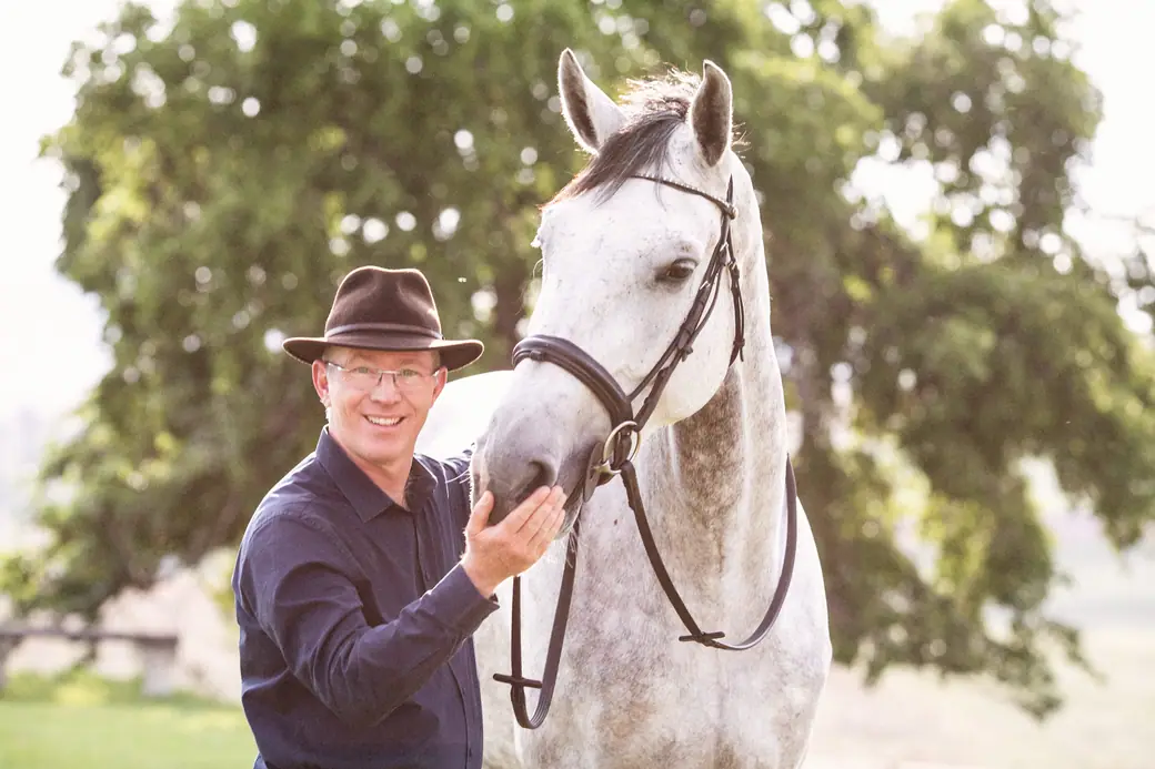 Jenku Dietrichsen, in a wide-brimmed hat and glasses, standing beside a grey horse in a bridle with one hand resting on its cheek.